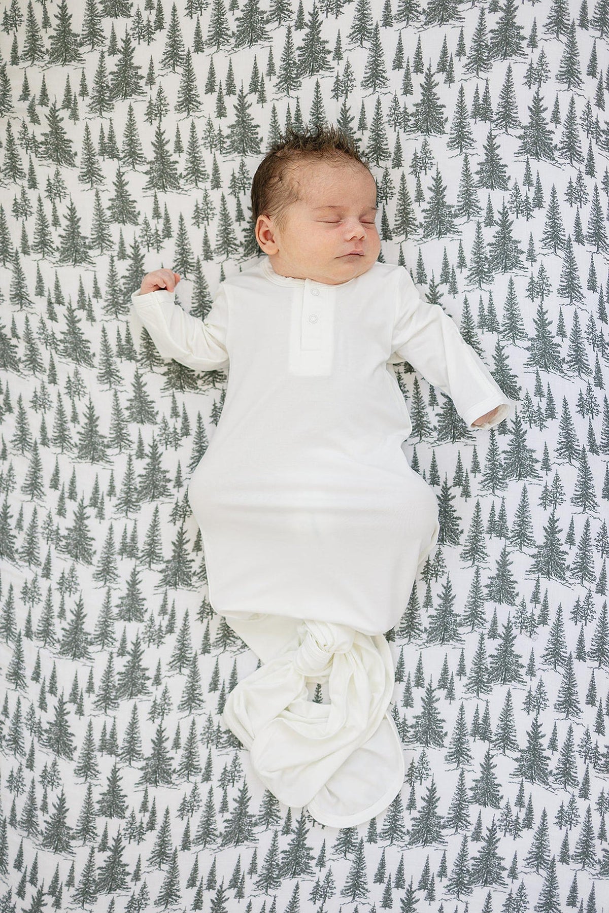 A sleeping baby in a white outfit rests on a crib sheet with a green tree pattern.