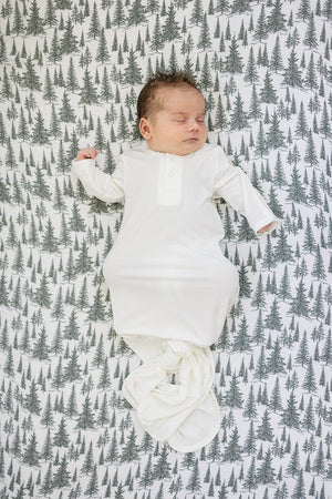 A sleeping baby in a white outfit rests on a crib sheet with a green tree pattern.