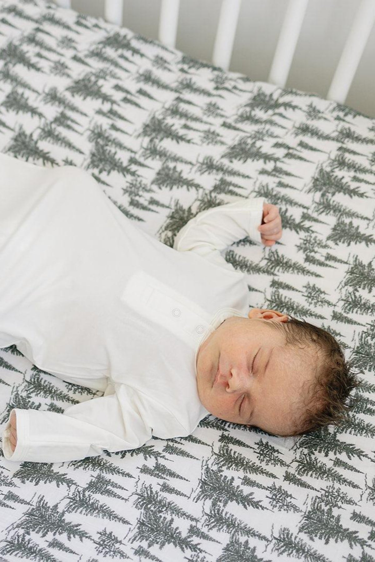 A baby in a white outfit sleeps on a crib sheet with a black tree pattern.