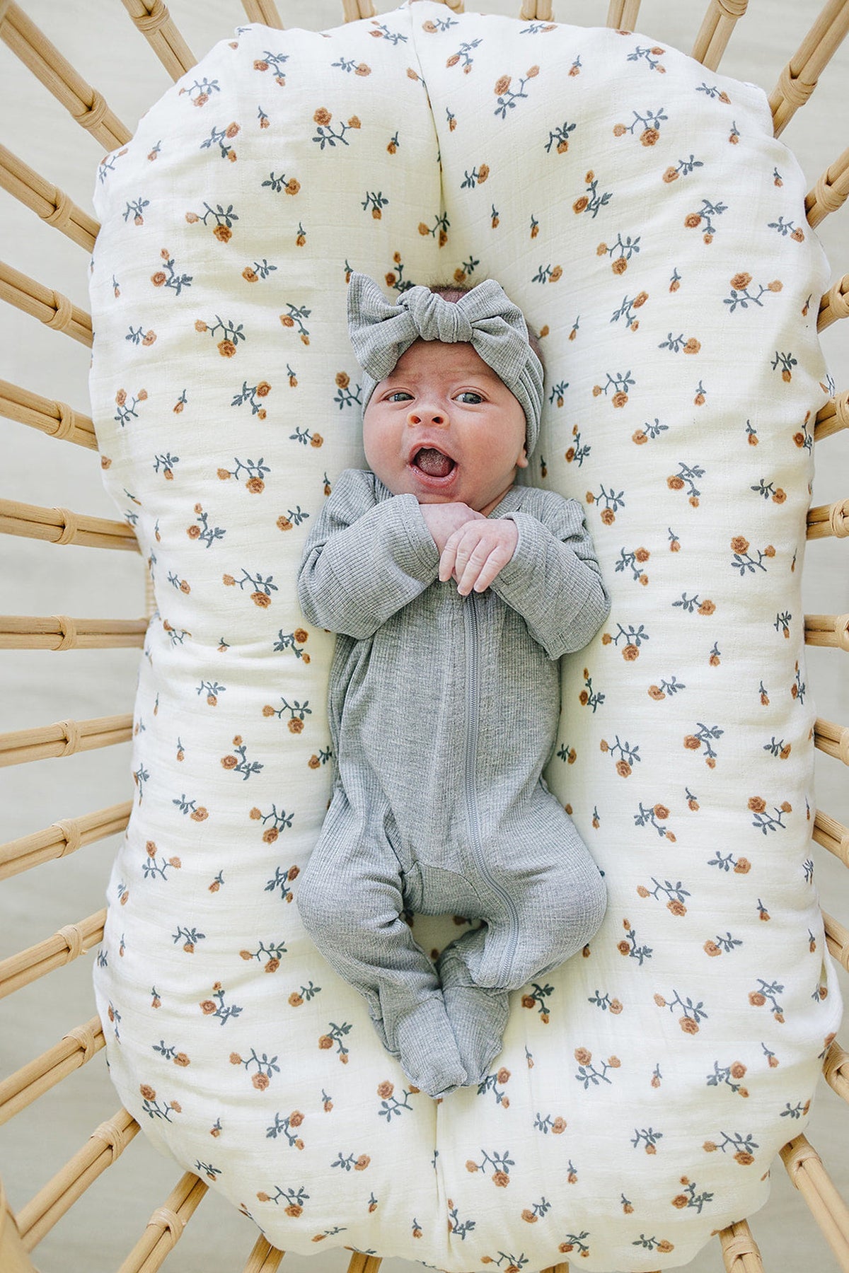 A baby wearing a grey ribbed head wrap and onesie lies on a floral-patterned cushion in a wicker bassinet.