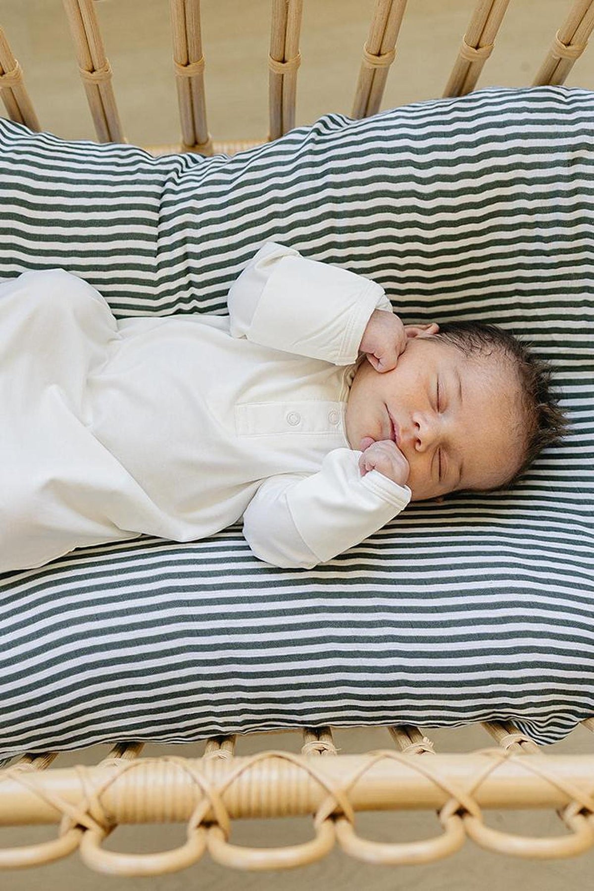 A baby sleeps in a white gown on a striped cushion in a wicker bassinet.