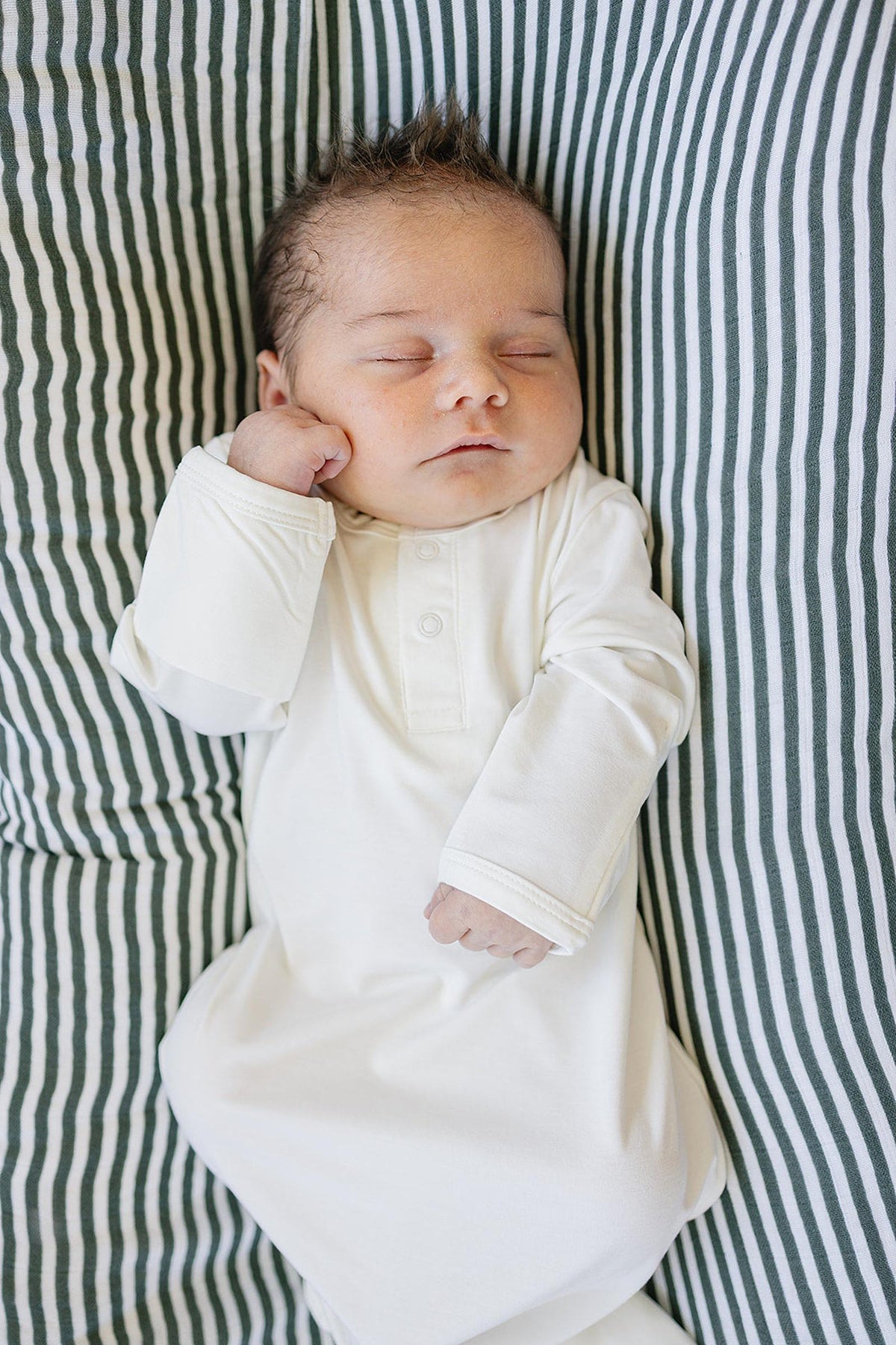 Newborn sleeping in a white gown on a striped cushion. The gown is soft and perfect for infants.