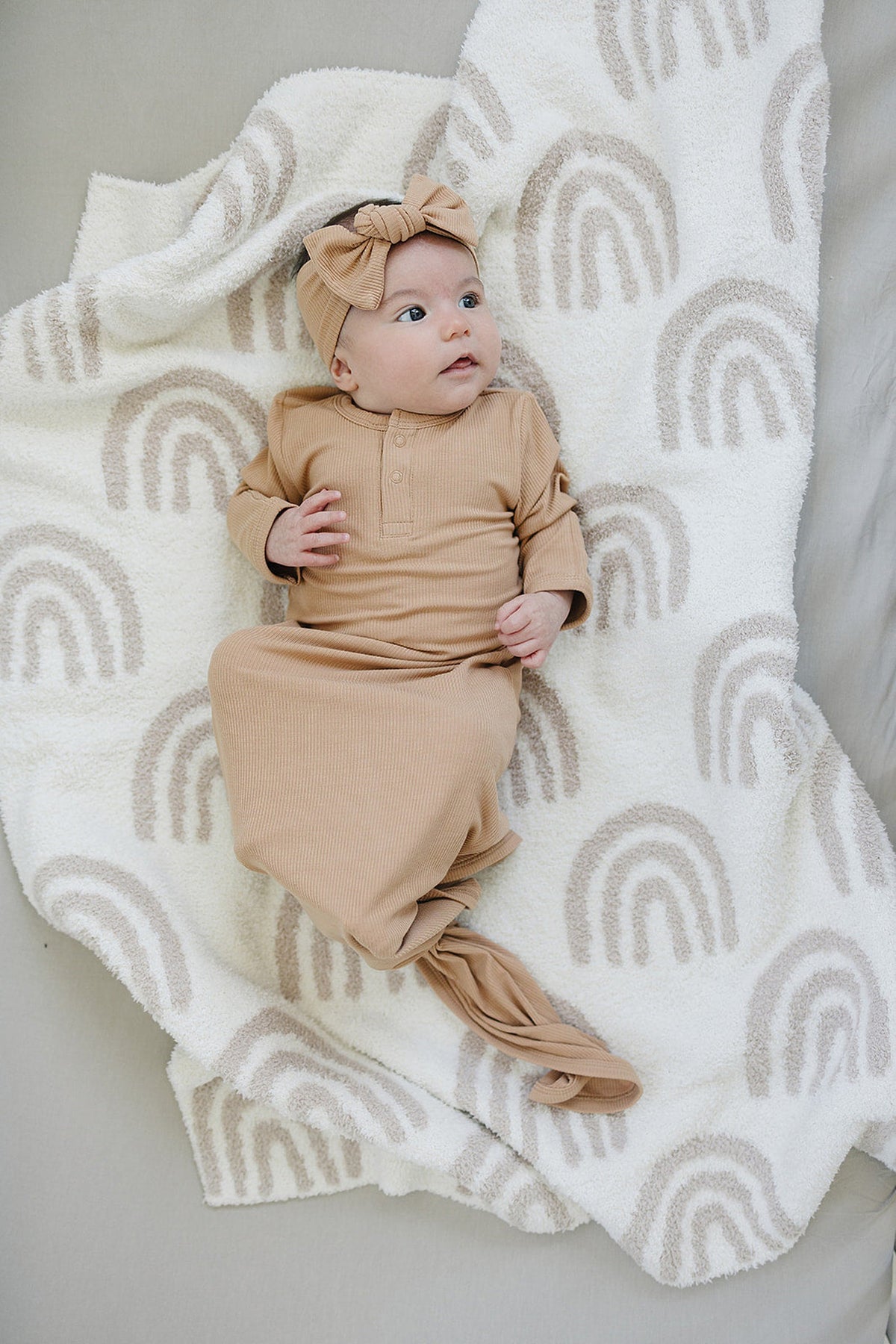 A baby wearing a latte-colored ribbed gown and head wrap lies on a blanket with beige rainbow patterns.