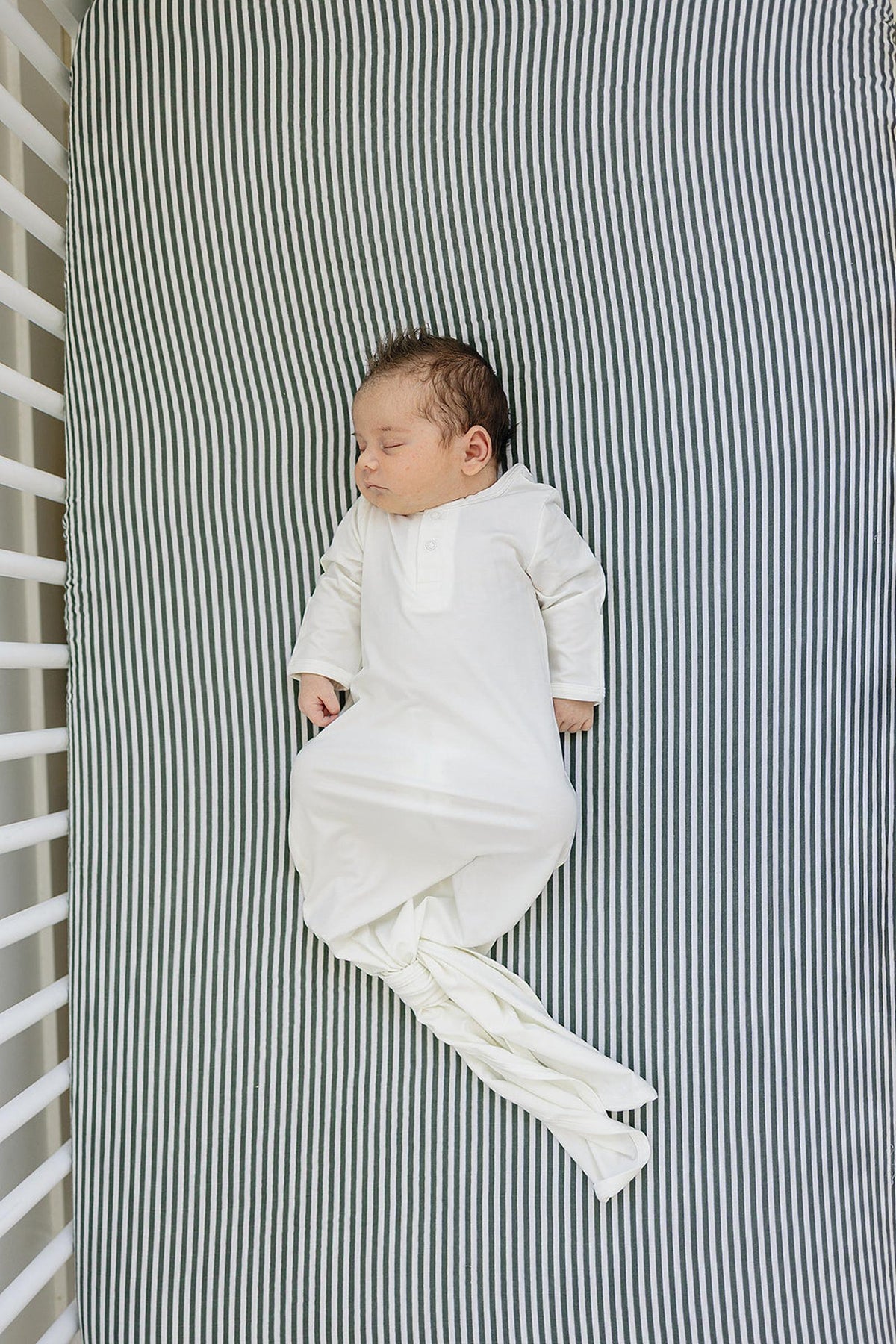 A baby sleeps peacefully on a green striped crib sheet, wearing a white sleep gown.