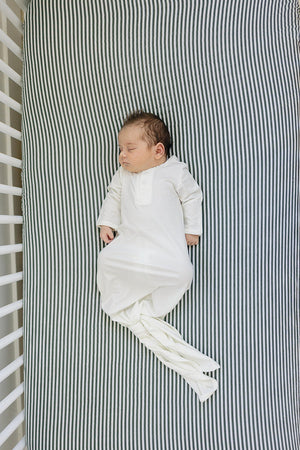 A baby sleeps peacefully on a green striped crib sheet, wearing a white sleep gown.