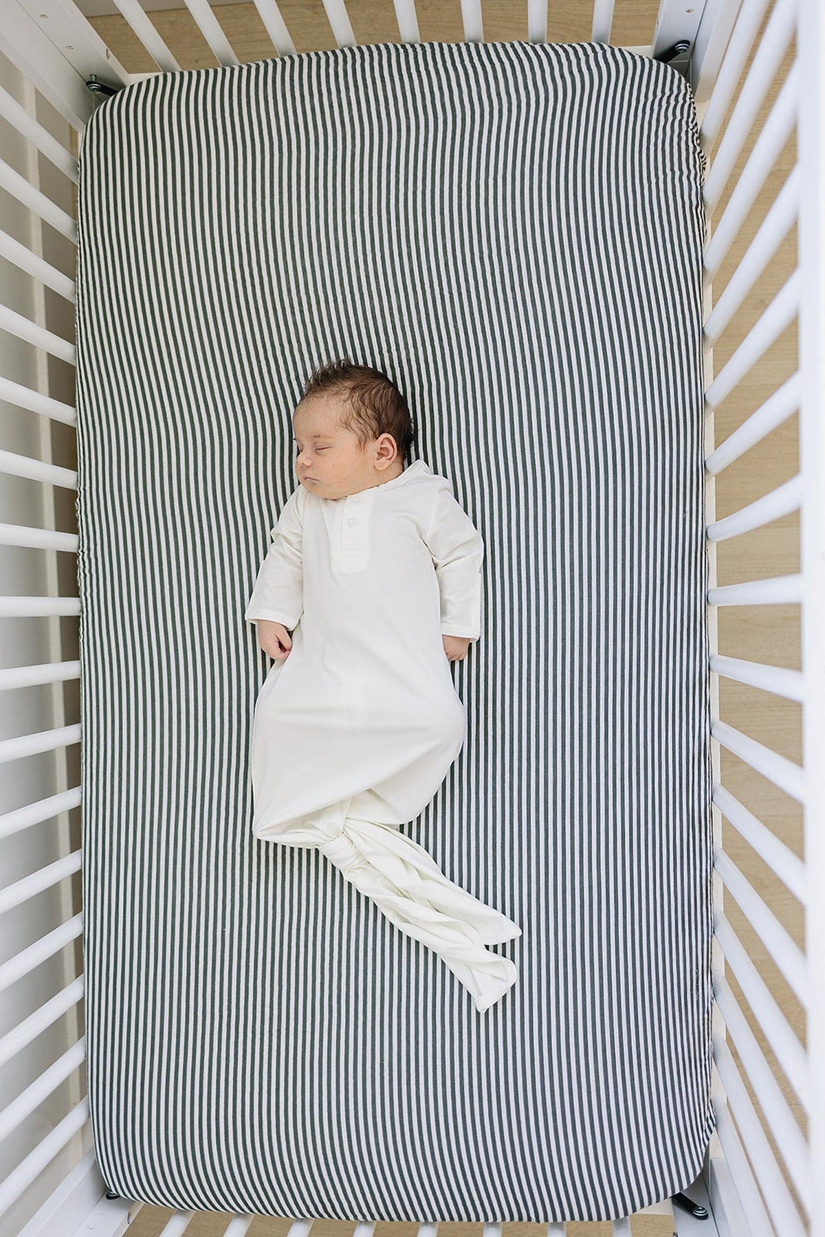 A baby sleeps in a crib wearing a white knot gown on a striped mattress.