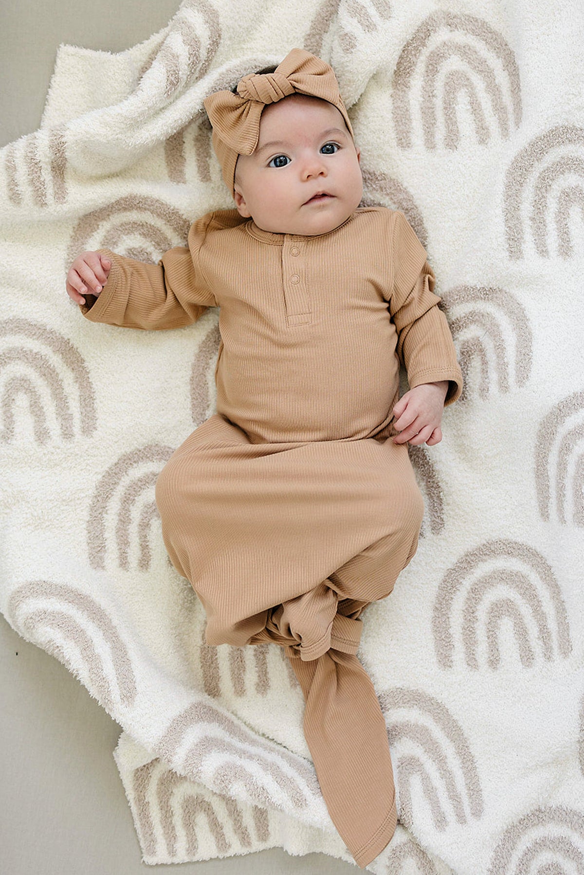 A baby lies on a soft, cream blanket with rainbow patterns, wearing a tan ribbed knot gown and a matching bow headband. The outfit is made from breathable bamboo fabric.