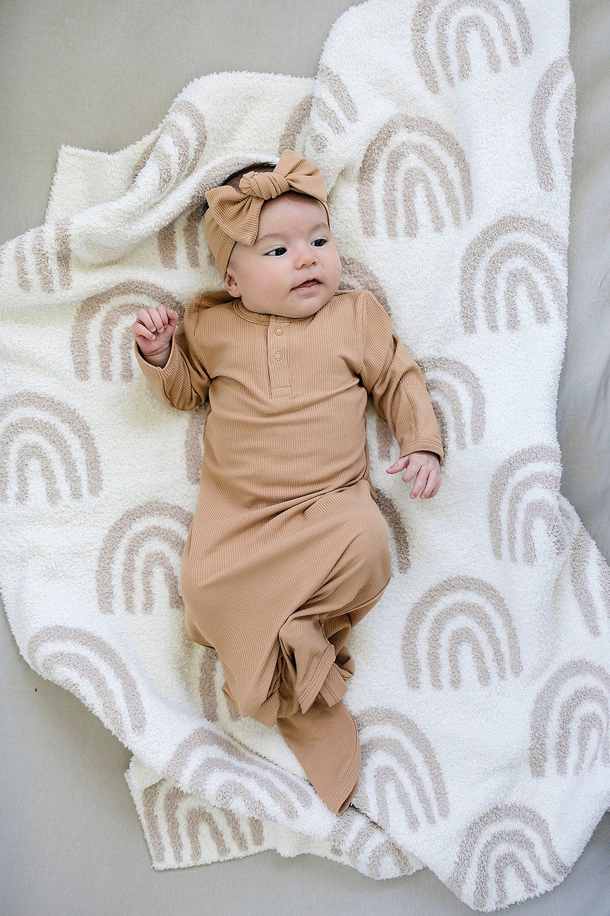 A baby lies on a soft, white blanket with a beige rainbow pattern, wearing a tan ribbed knot gown and a matching headband.