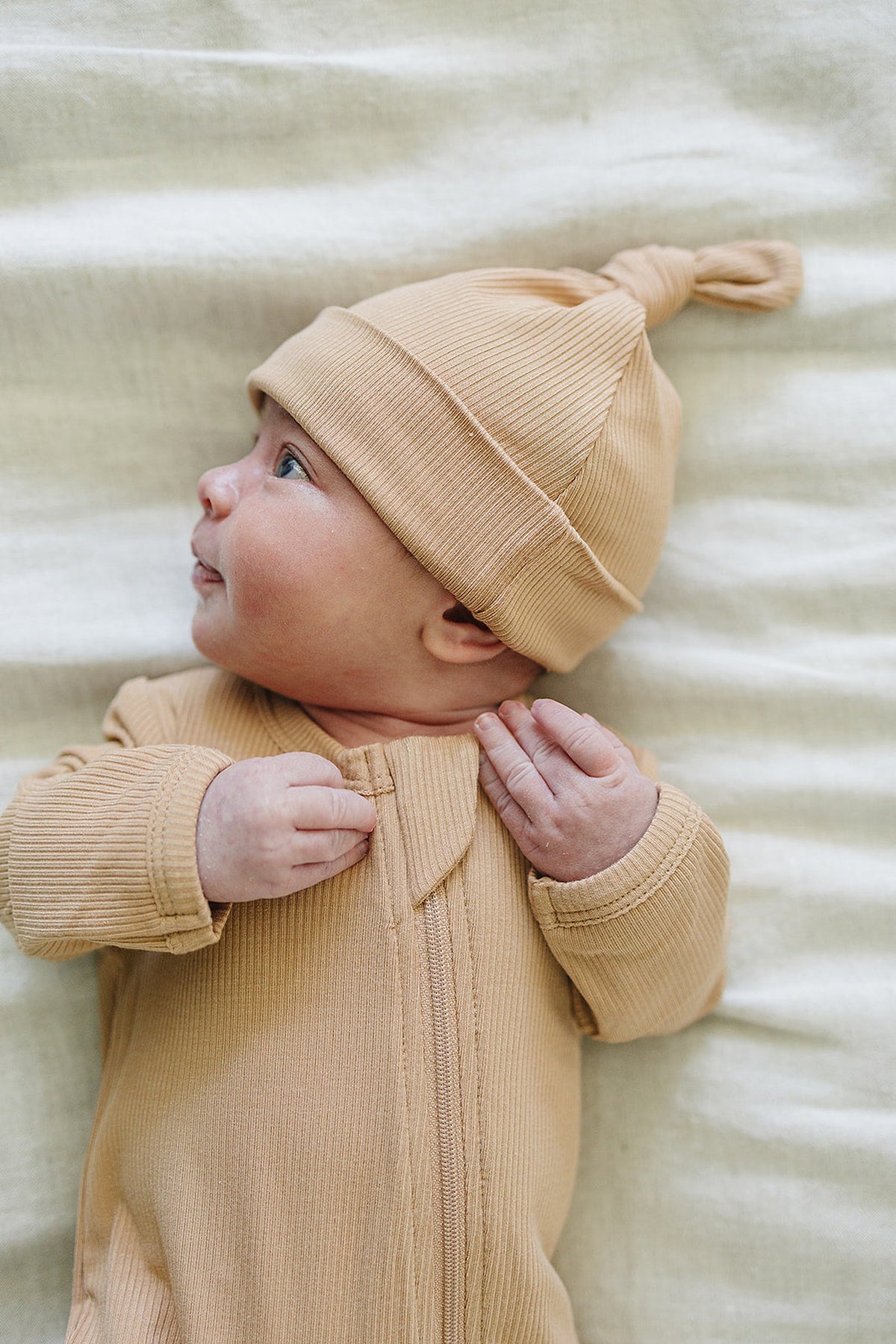 A baby wearing a latte-colored, ribbed bamboo knot hat and matching outfit lies on a soft, light blanket.
