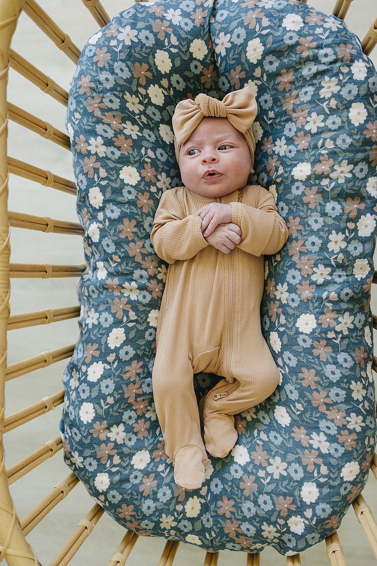 A baby wearing a latte-colored ribbed outfit and head wrap lies on a floral-patterned cushion.