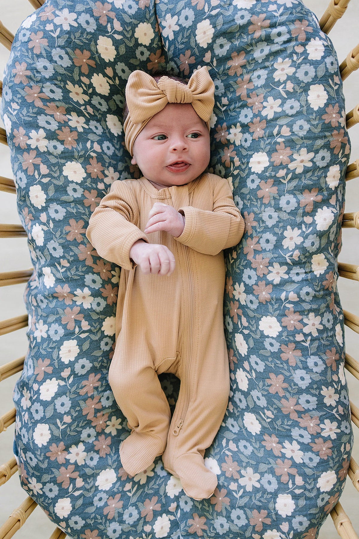 Baby wearing a latte-colored ribbed zipper pajama and matching headband, lying on a floral-patterned cushion.
