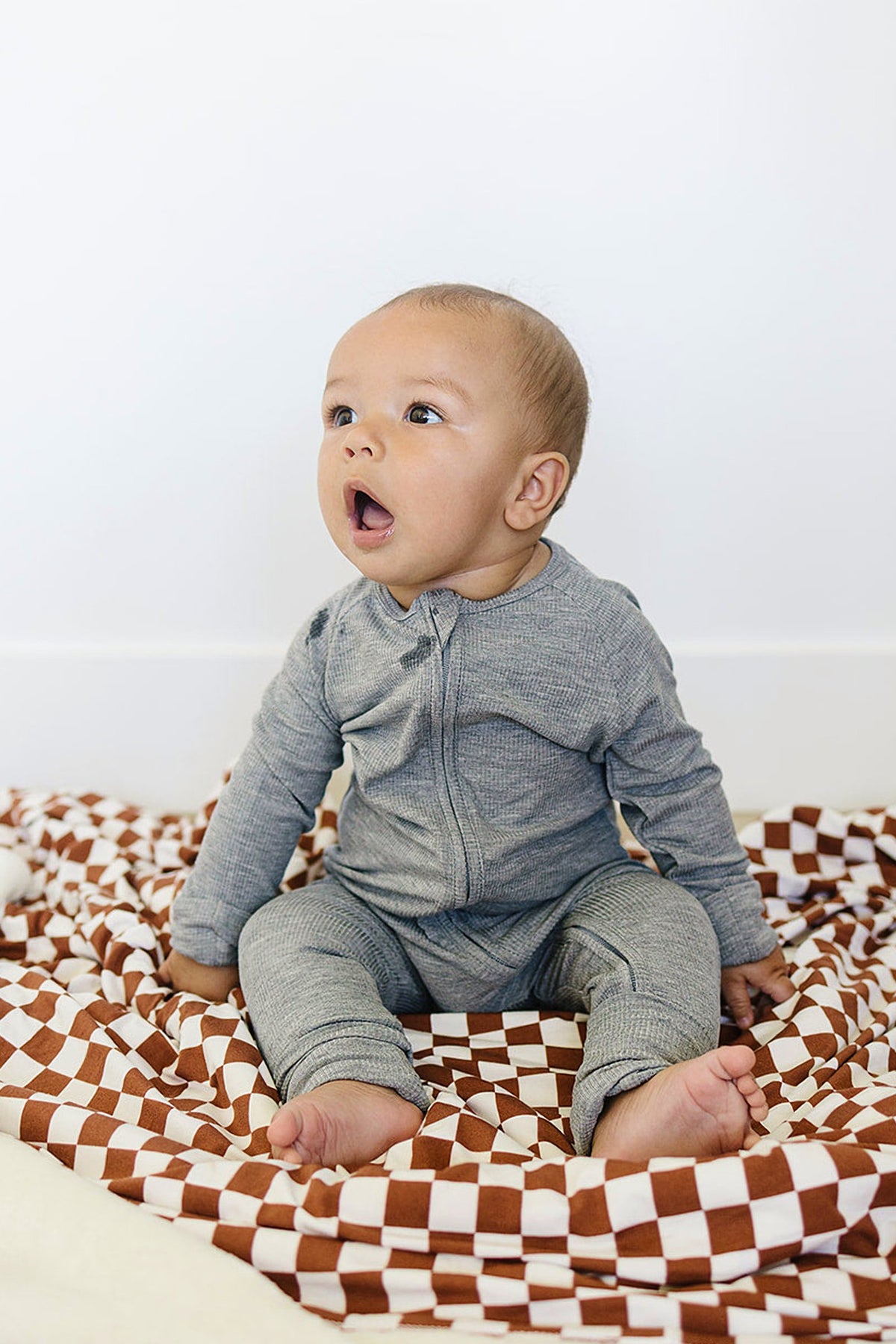 Baby wearing a heather grey ribbed zipper pajama sits on a checkered brown and white blanket, looking curious.