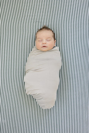 A baby swaddled in a beige blanket lies on a green striped crib sheet, peacefully asleep.
