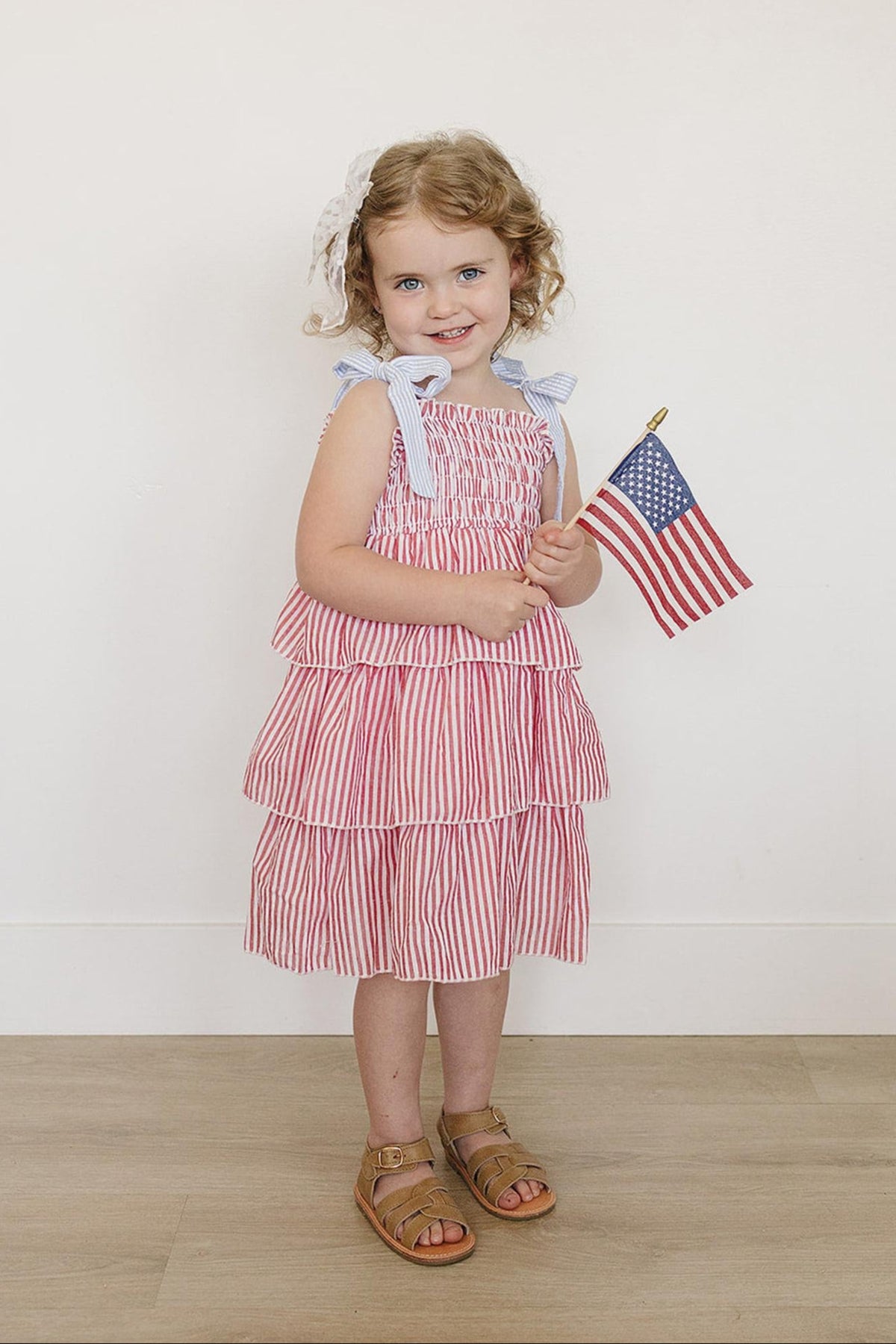 A young child in a red striped ruffle dress holds an American flag, standing on a wooden floor.
