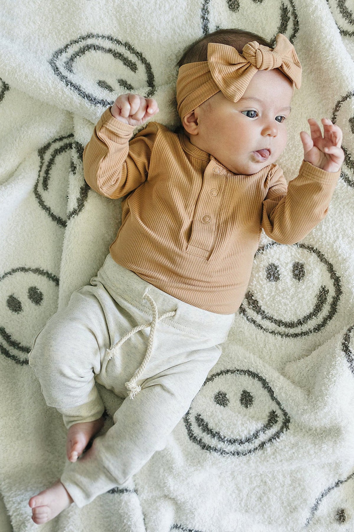 Baby wearing a brown ribbed long sleeve bodysuit and headband, lying on a white blanket with smiley faces.