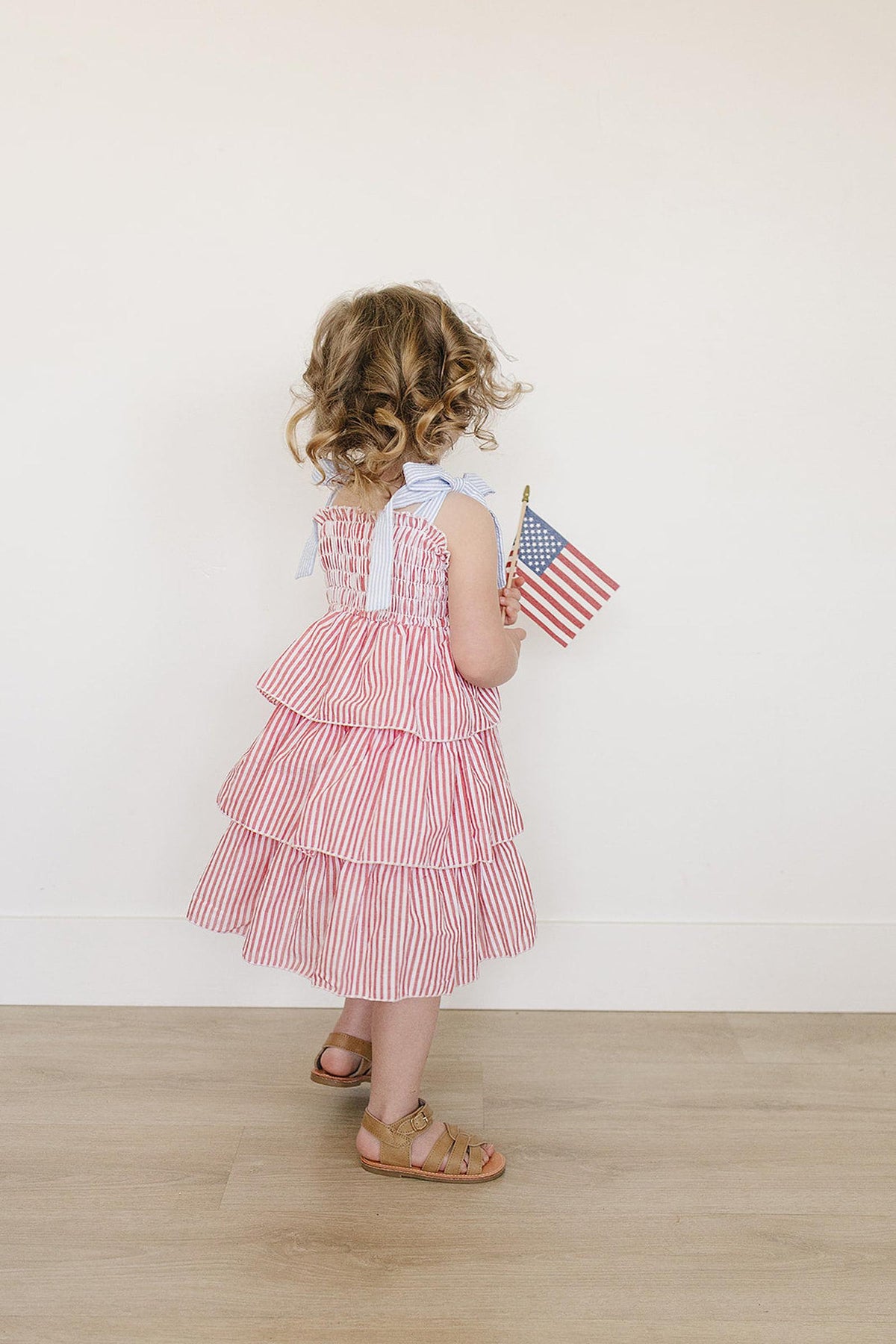 A toddler in a red striped ruffle dress holds an American flag, standing on a wooden floor.