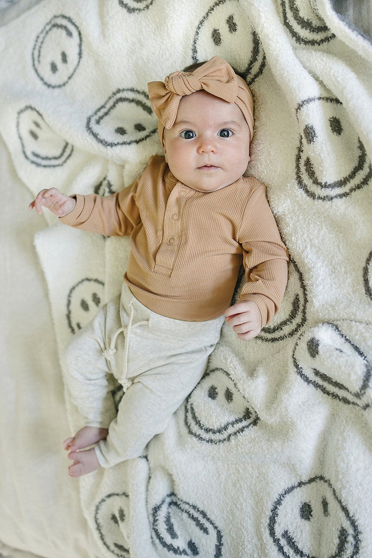 A baby wearing a ribbed tan bodysuit and bow headband lies on a blanket with smiley faces.