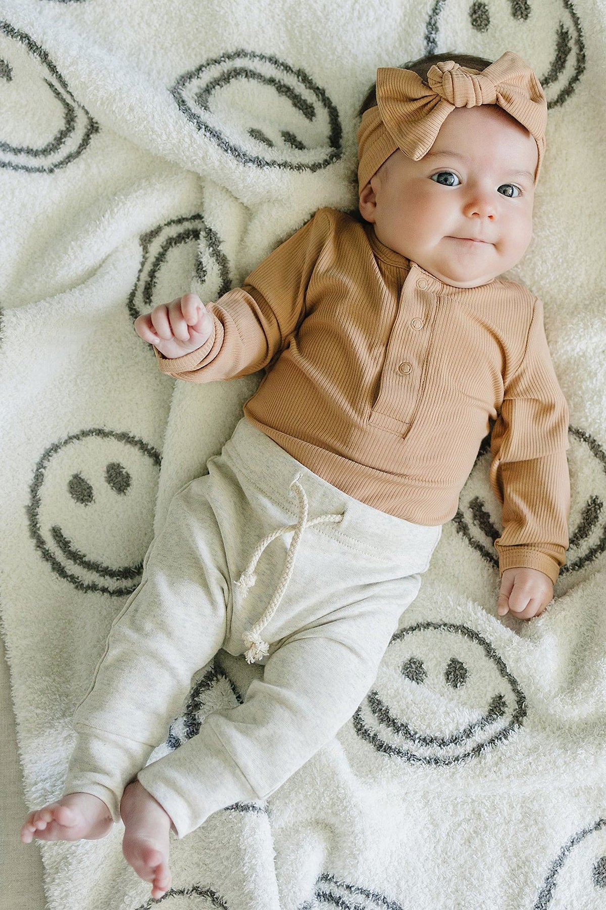 A baby wearing a latte-colored ribbed bodysuit and headband lies on a blanket with smiley faces.