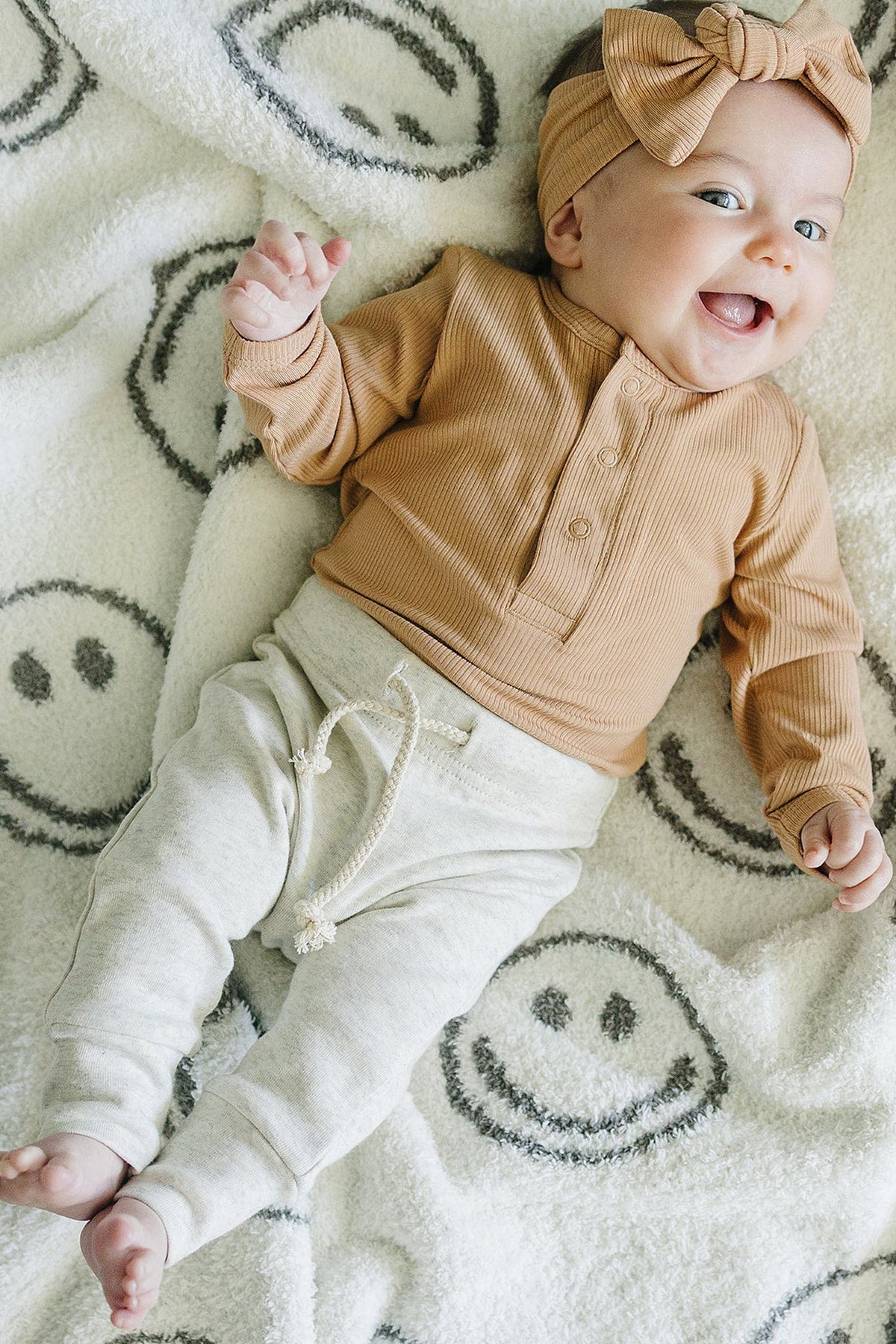 A baby wearing a latte ribbed head wrap and matching top lies on a smiley-face patterned blanket, smiling.