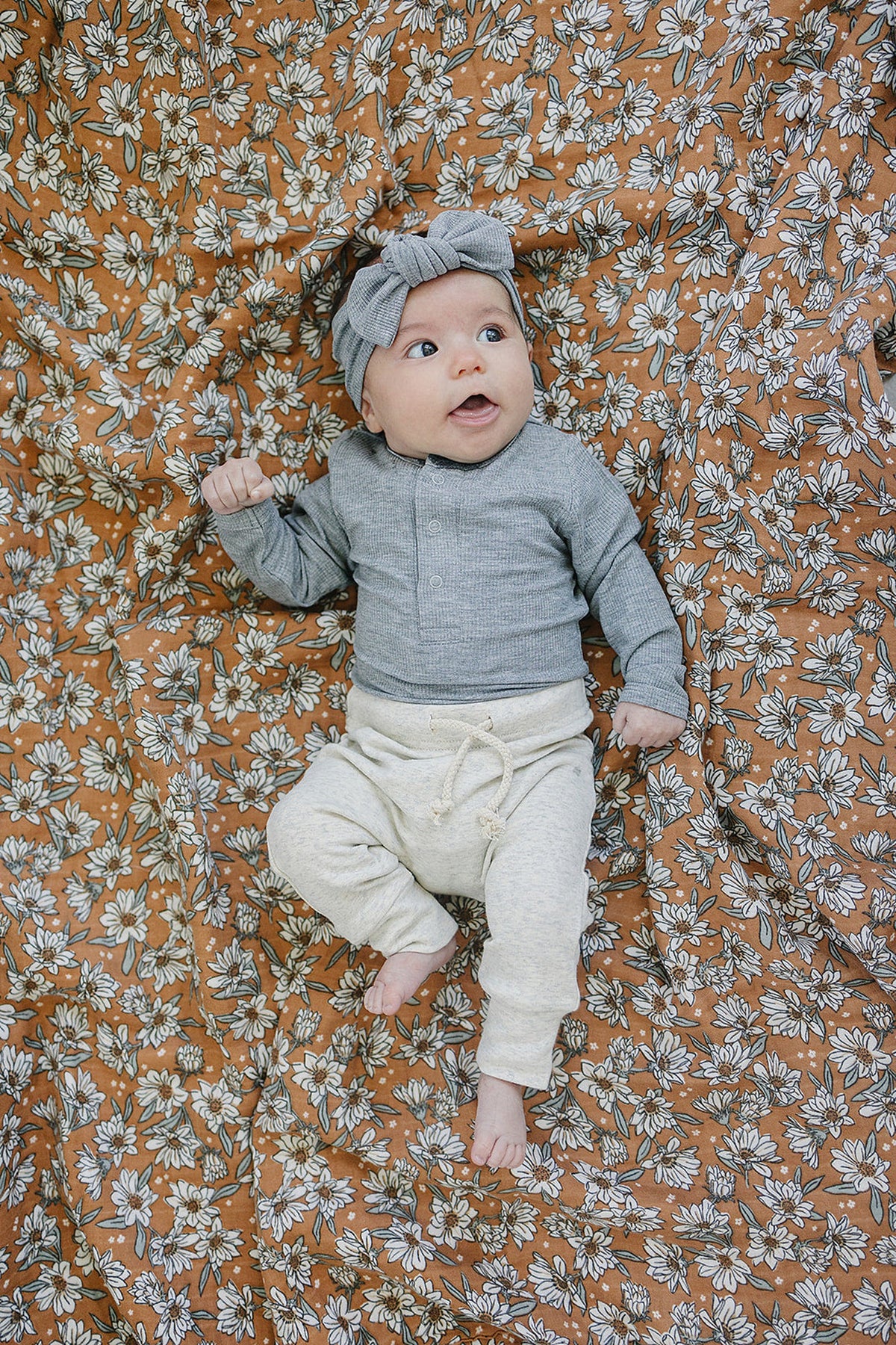 Baby wearing a grey ribbed head wrap and outfit, lying on a floral-patterned blanket.