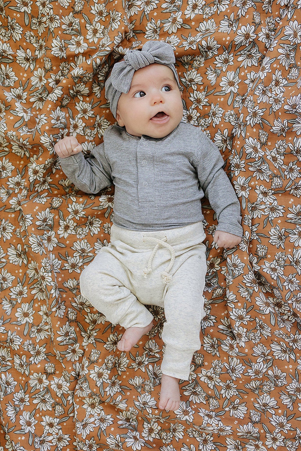 A baby in a grey ribbed bodysuit and headband lies on a floral-patterned blanket.