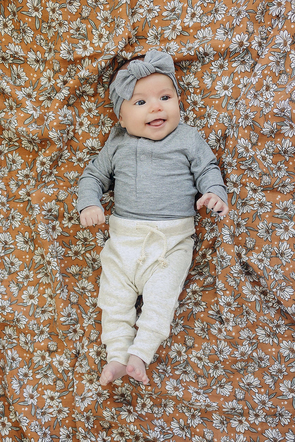 A baby in a grey ribbed bodysuit and cream pants lies on a floral-patterned blanket, smiling.
