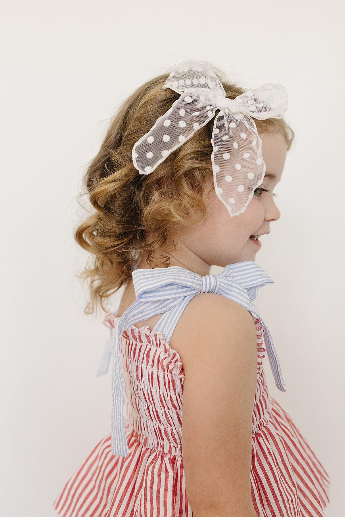 A young girl in a red striped dress with ruffle details, wearing a polka dot bow headband.