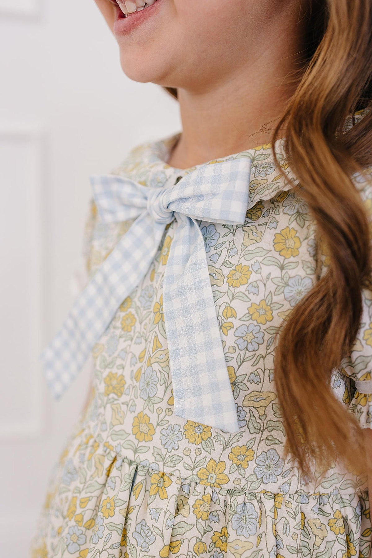 A young girl smiles while wearing a floral dress featuring a light blue gingham bow. The dress showcases a pastel floral pattern, perfect for springtime occasions.