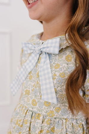 A young girl smiles while wearing a floral dress featuring a light blue gingham bow. The dress showcases a pastel floral pattern, perfect for springtime occasions.