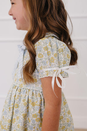 A young girl models a sunny floral dress with a classic Peter Pan collar, featuring pastel yellow and blue flowers.