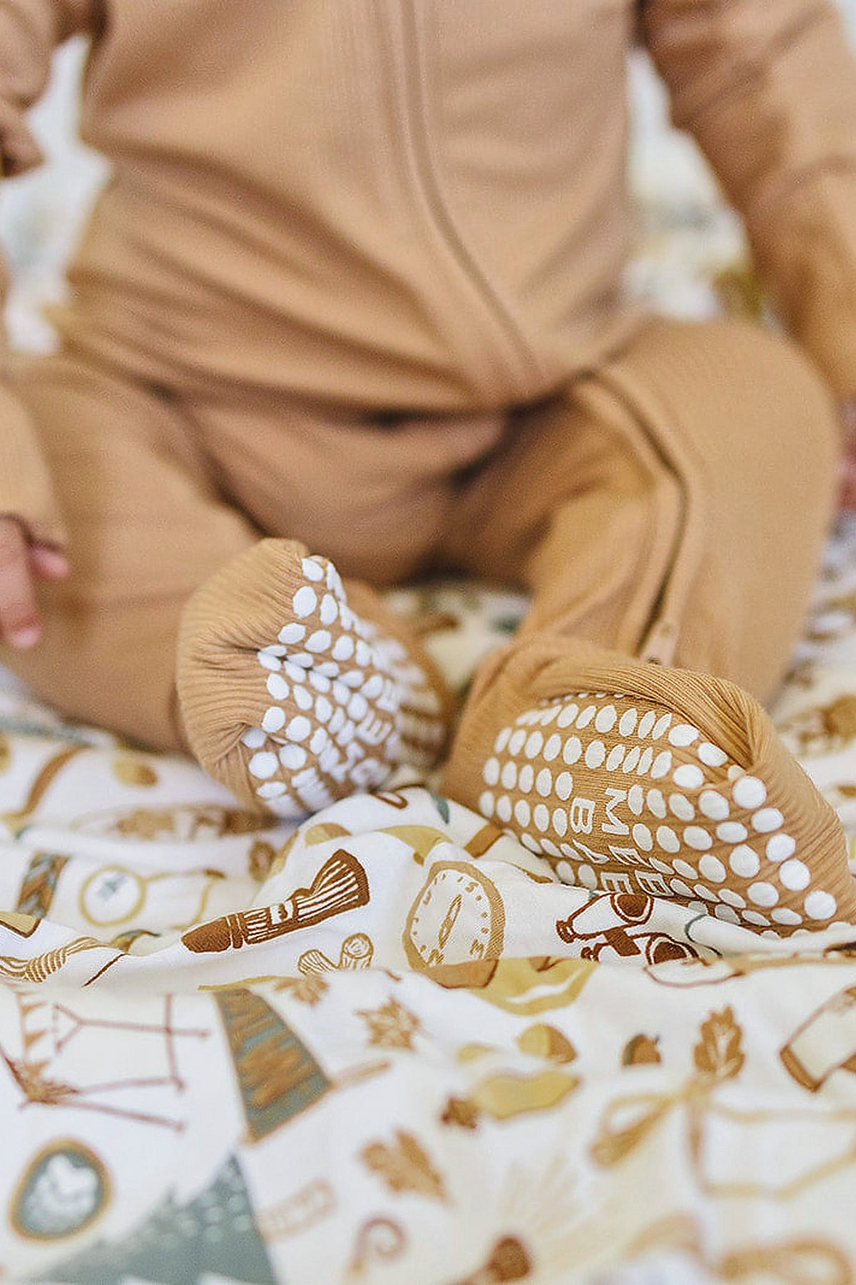 A baby wearing a latte-colored ribbed zipper pajama with textured foot grips sits on a patterned blanket.
