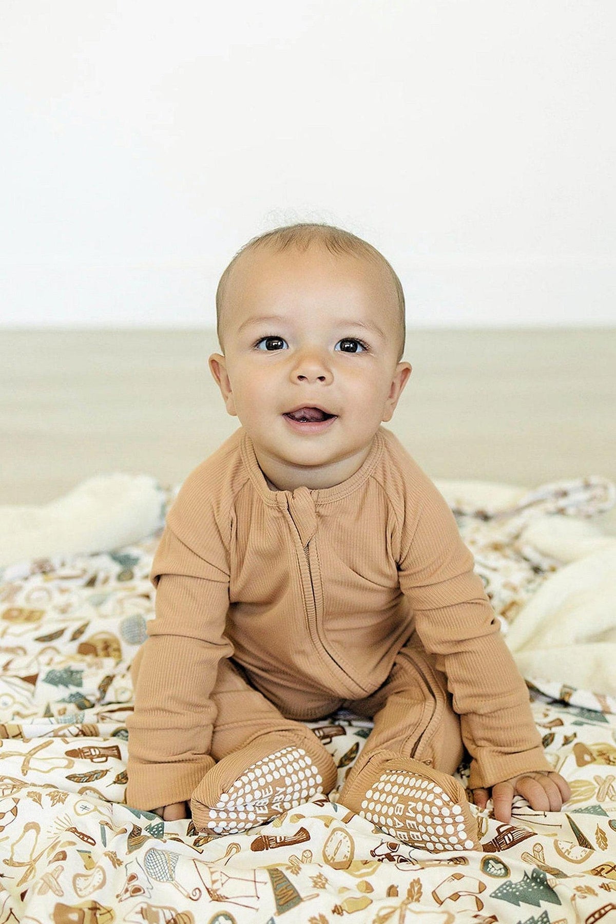 A baby in a latte-colored ribbed zipper pajama sits on a patterned blanket, smiling warmly.