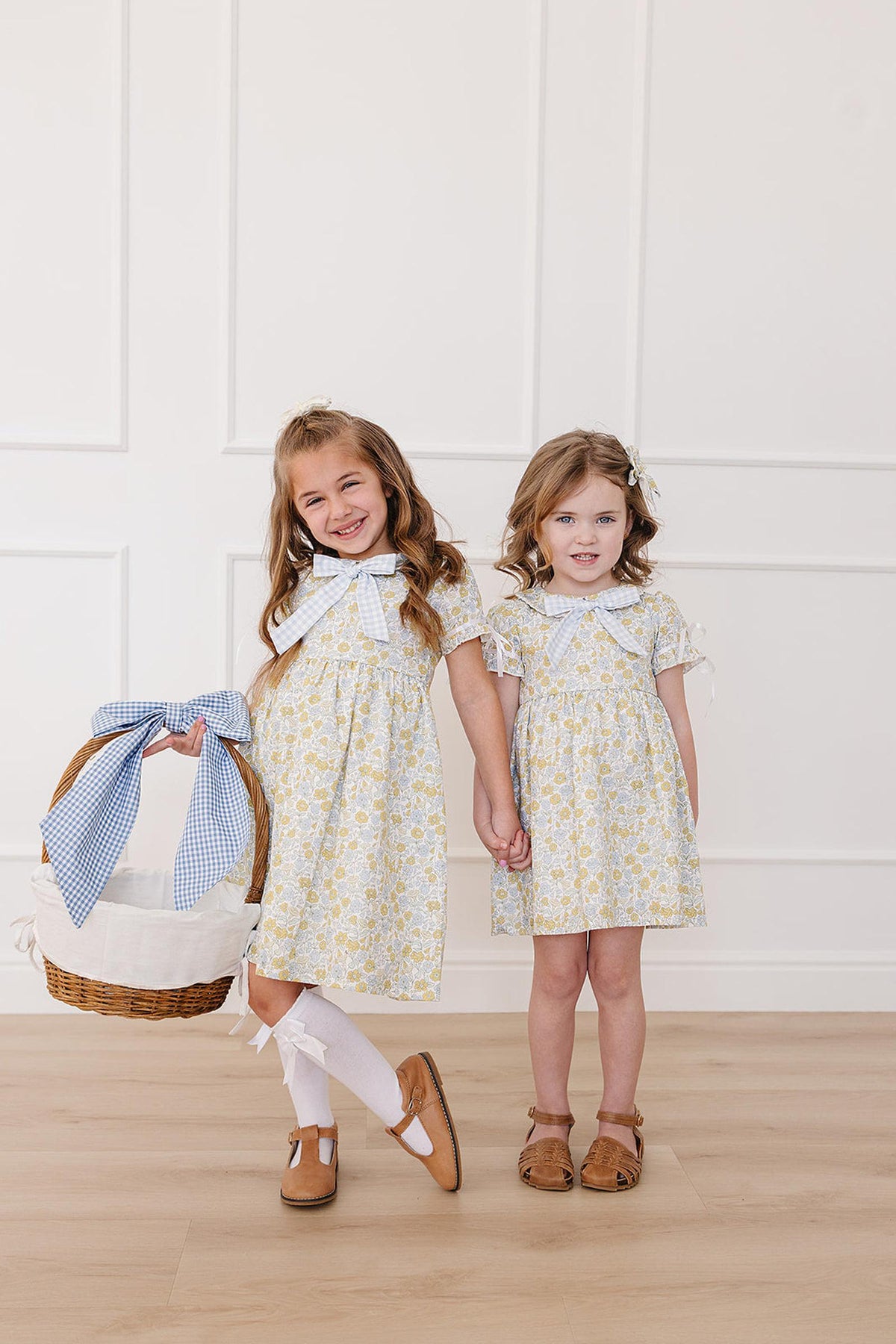 Two young girls stand side by side, wearing matching sunny floral dresses with Peter Pan collars.