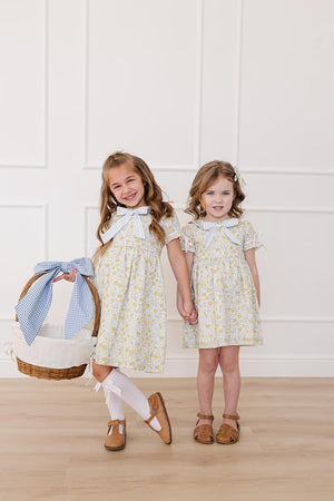 Two young girls stand side by side, wearing matching sunny floral dresses with Peter Pan collars.