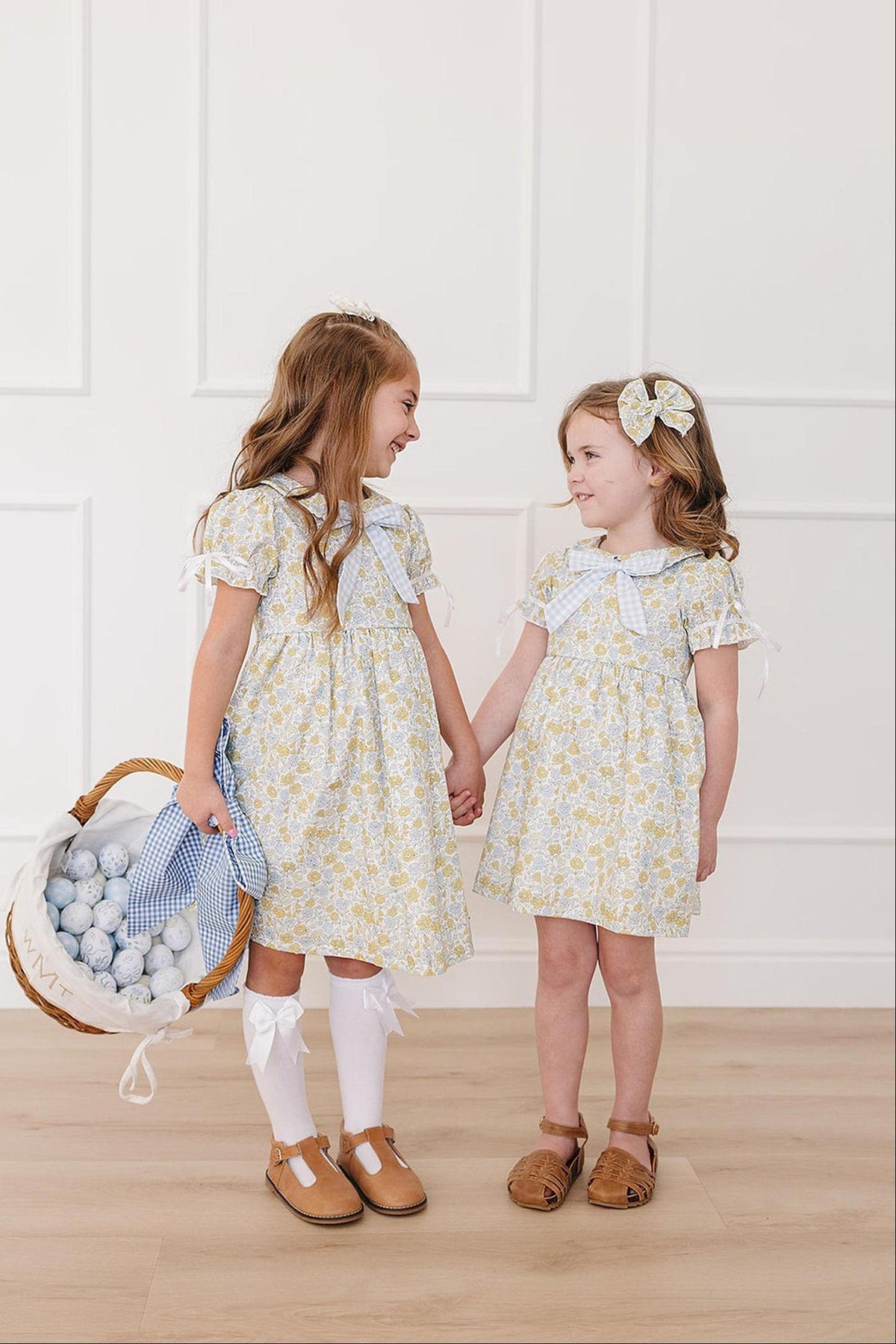 Two young girls hold hands, wearing matching sunny floral dresses with Peter Pan collars and bows. The setting features a light, airy background, enhancing the dresses cheerful, vintage style.