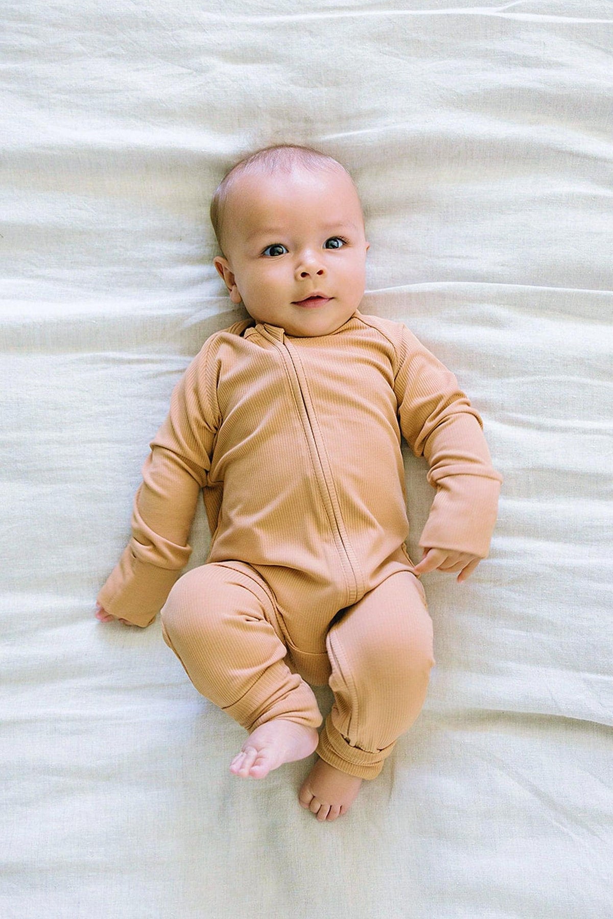 A baby in a latte-colored, ribbed bamboo zipper pajama lies on a soft, light-colored blanket.