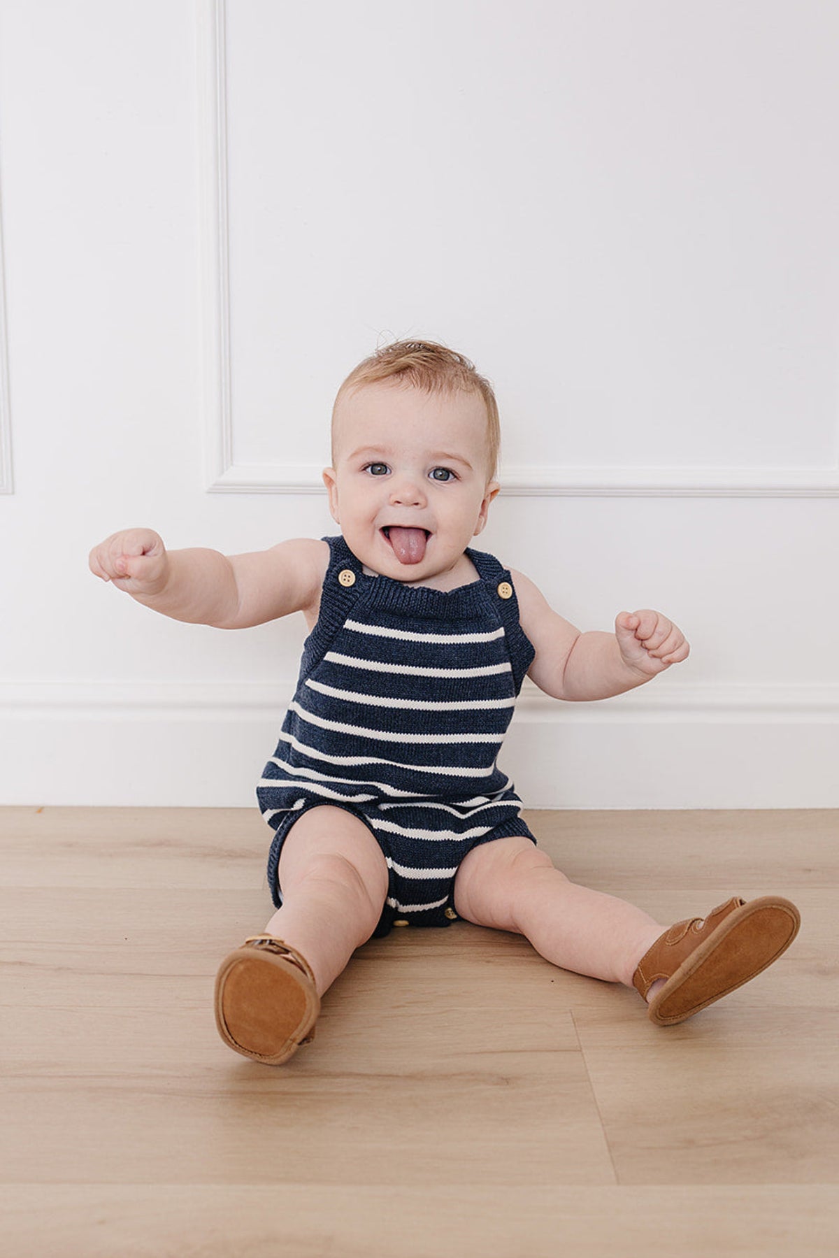 A baby sits on a wooden floor, wearing a navy and white striped knit bubble romper with snap closures. The child playfully sticks out their tongue, showcasing a joyful expression.