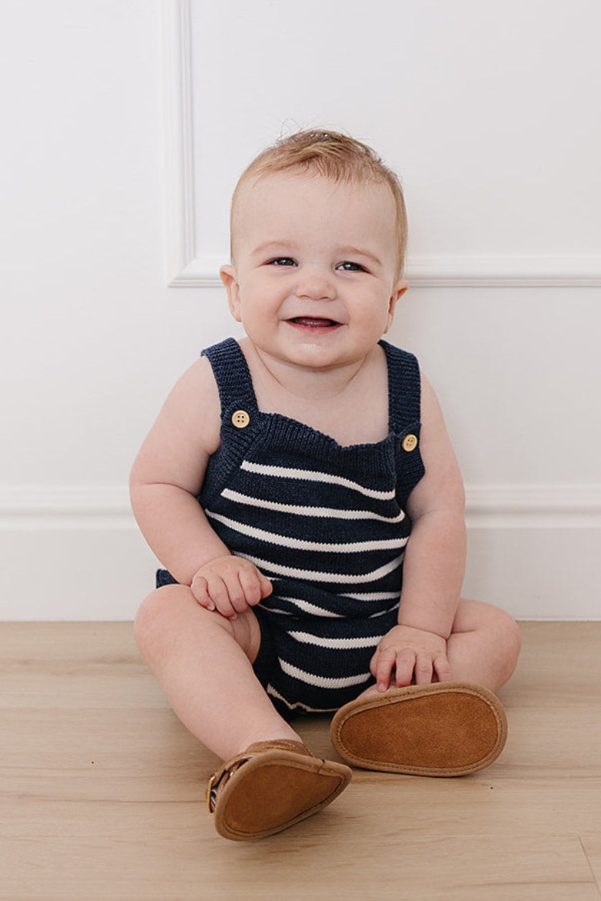 A happy baby sits on a light wooden floor, wearing a navy and white striped knit bubble romper with wooden button details.