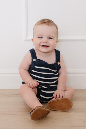 A happy baby sits on a light wooden floor, wearing a navy and white striped knit bubble romper with wooden button details.