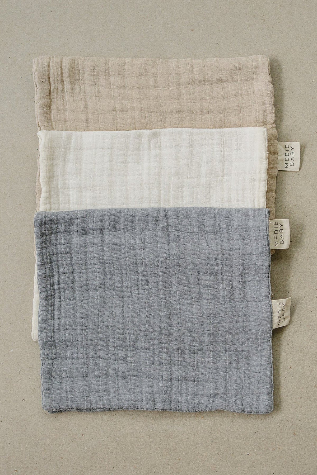 Three Mebie Baby muslin burp cloths in dusty blue, cream, and beige with visible tags on beige background.