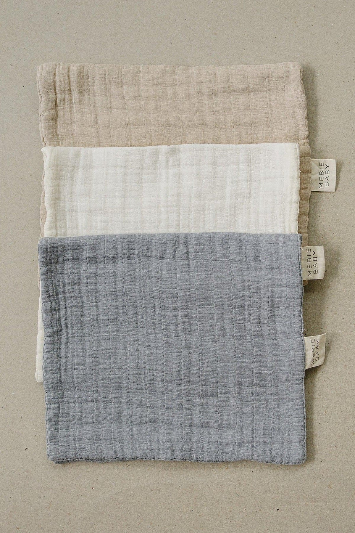 Three muslin burp cloths in oatmeal, cream, and gray colors are neatly stacked on a light background, showcasing their soft.