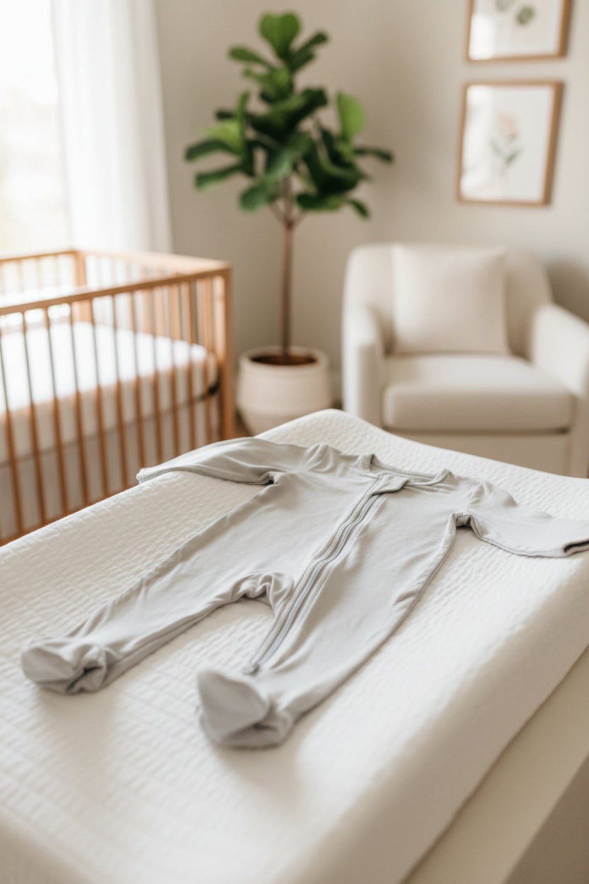 Gray baby footed pajama with zipper on changing table in a nursery with a crib, chair, and plant.