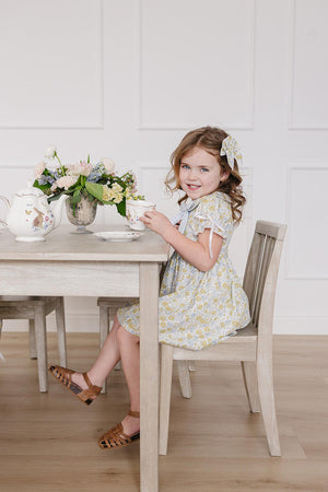 A young girl sits at a light wooden table, wearing a floral dress with a Peter Pan collar and bows on the sleeves, enjoying tea beside a vase of flowers.