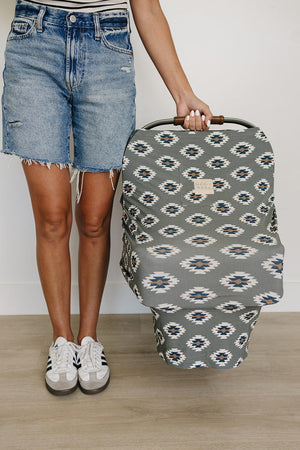 A person holds a Mebie Baby car seat cover with a Southwest Aztec pattern, wearing denim shorts and sneakers.