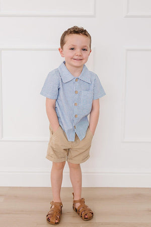 A young boy stands in a light, airy room wearing a blue and white striped button-up shirt with a chest pocket, paired with beige shorts and brown sandals.