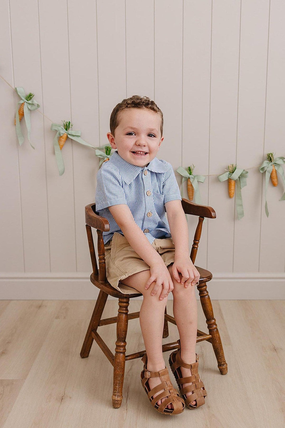 A young boy sits on a wooden chair, wearing a blue-striped button-up shirt and beige shorts. The background features a decorative garland with carrots, creating a cheerful atmosphere.