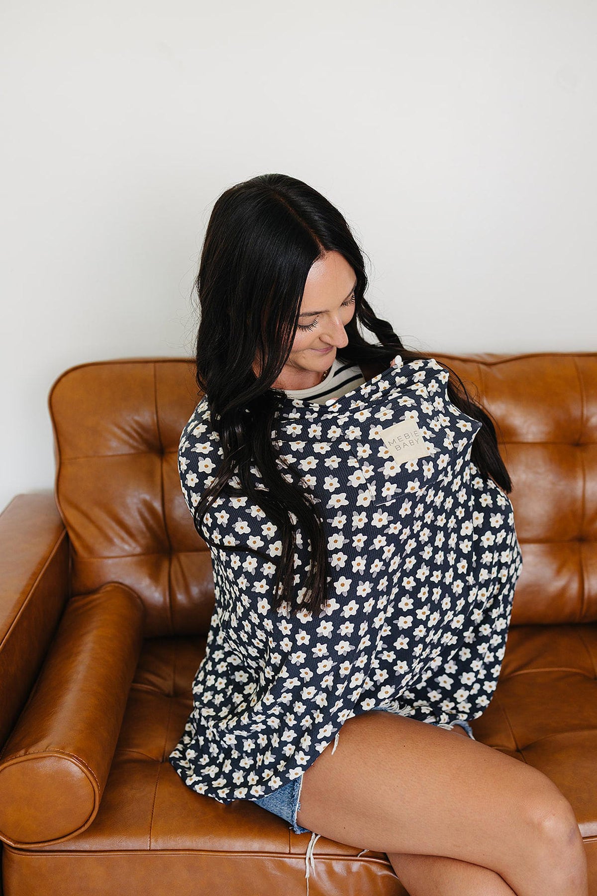 A woman sits on a brown leather couch, using a navy daisy-patterned Mebie Baby cover.