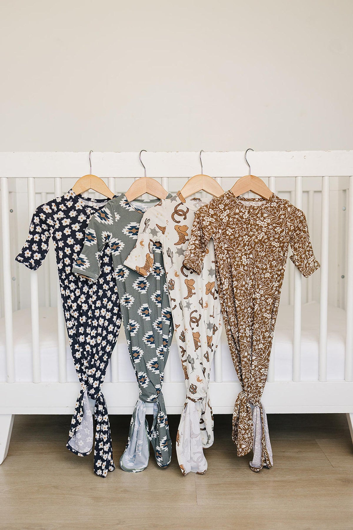 Four patterned baby gowns hang on wooden hangers in front of a white crib, showcasing floral and cowboy designs.