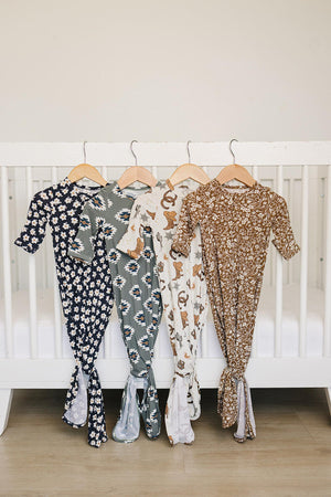 Four patterned baby gowns hang on wooden hangers in front of a white crib, showcasing floral and cowboy designs.