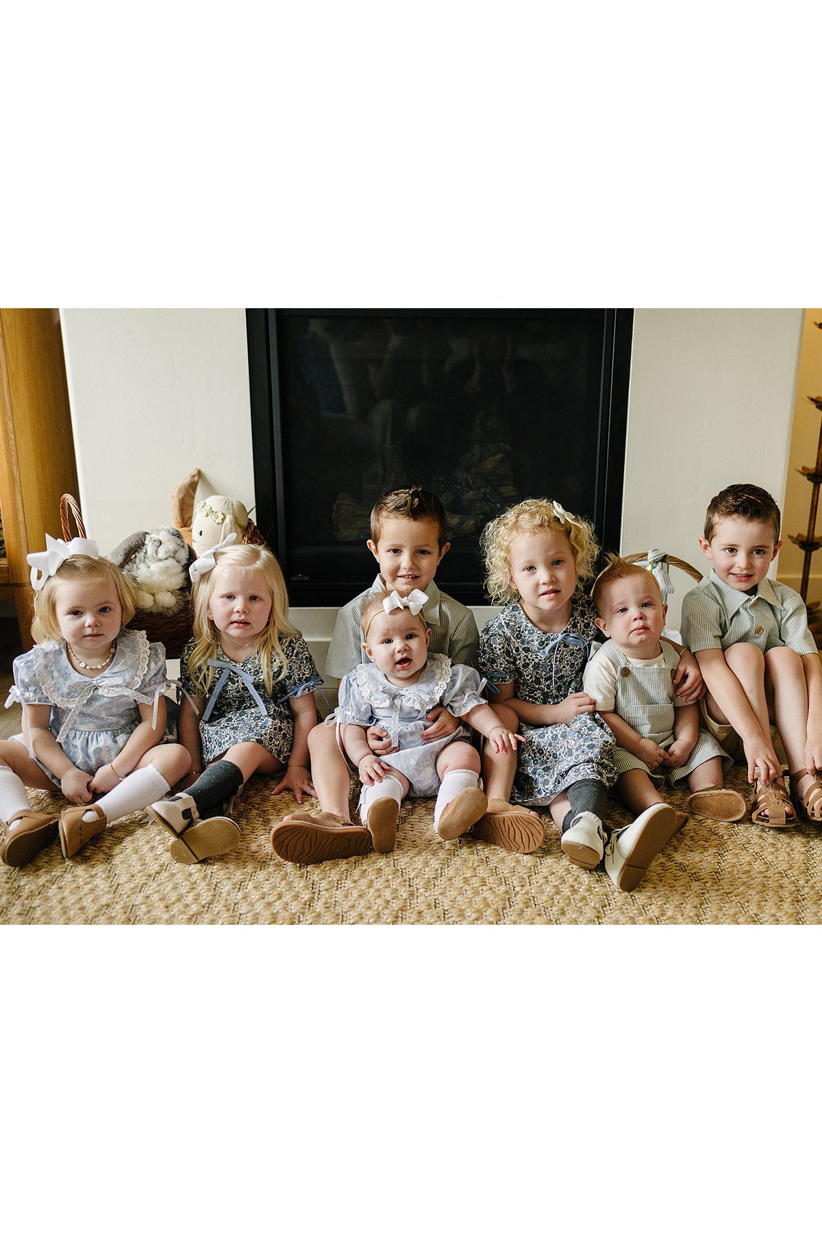 Seven children in coordinated outfits sit on a woven rug in front of a fireplace, smiling and posing.